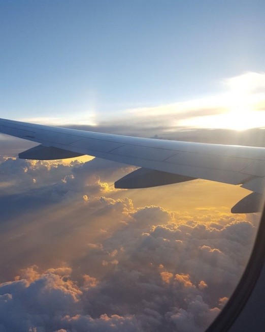 Photo take from an airplane window, the sky is light yellow and blue and we can see the plane's wing